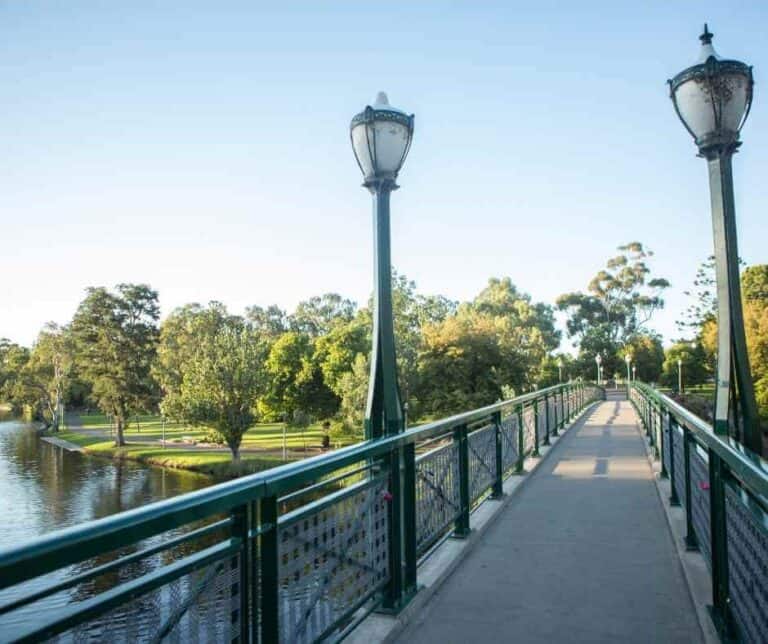 A pedestrian bridge over the River Torrens in Adelaide's Botanic Gardens, bathed in sunshine with verdant trees along the banks, indicating the mild weather conditions and answering the query, 'Does snow ever fall in Adelaide?'