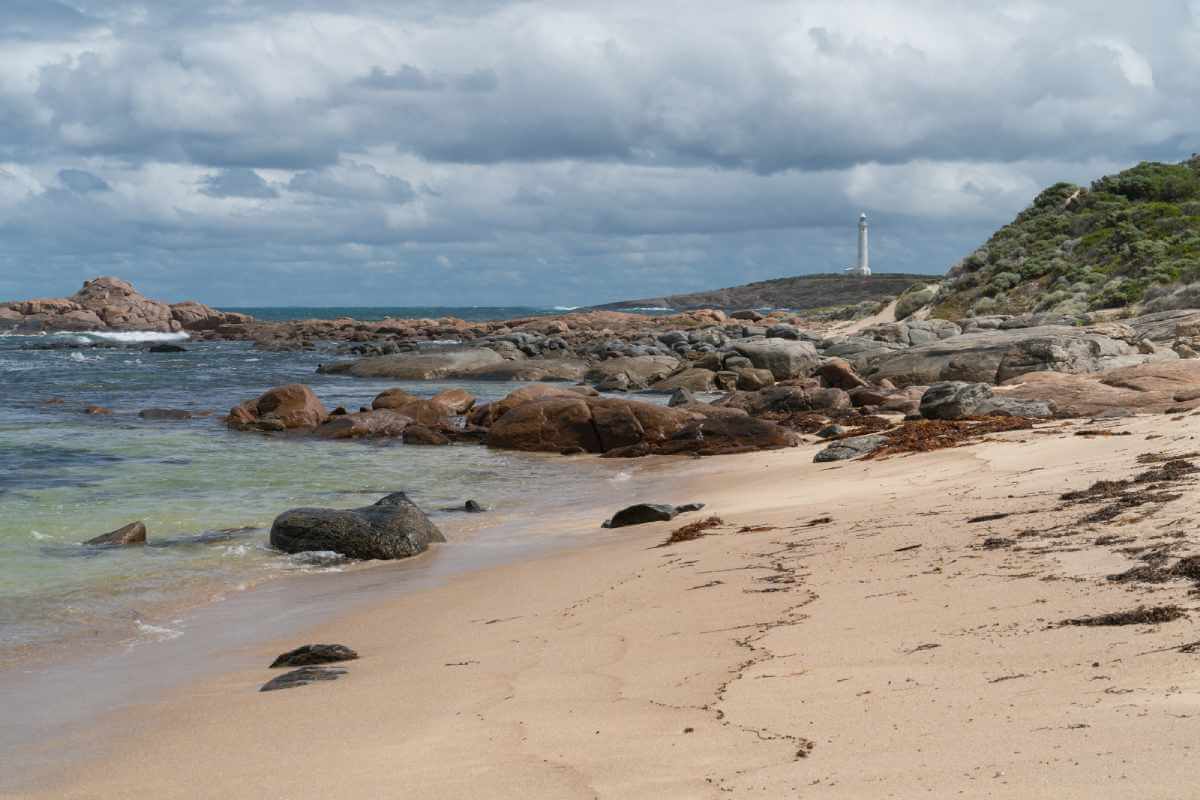 Calm winter beach with scattered rocks leading to Cape Leeuwin Lighthouse, illustrating tranquil things to do in Margaret River in winter, like coastal walks and lighthouse visits.