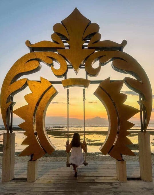 Kate enjoys a serene moment on the famous Ombak Sunset swing in the Gili Islands, silhouetted against a stunning sunset framed by an artistic golden arch. Swinging here is a must-do among the many Gili Islands activities, offering a perfect blend of adventure and relaxation.