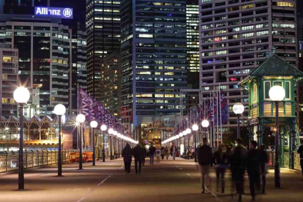 People walking along Pyrmont Bridge at night with the illuminated Sydney skyline in the background, a scenic route for evening walking tours in Sydney.
