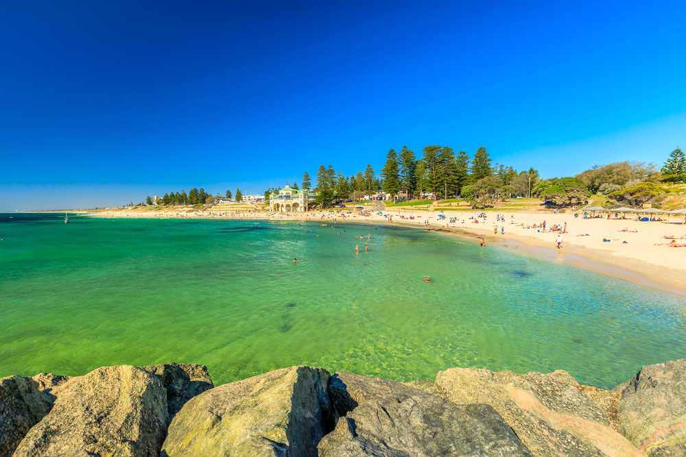 Clear turquoise waters and sunbathers at Cottesloe Beach, a family-friendly destination among the beaches in Western Australia, with the iconic Indiana Tea House in the background.
