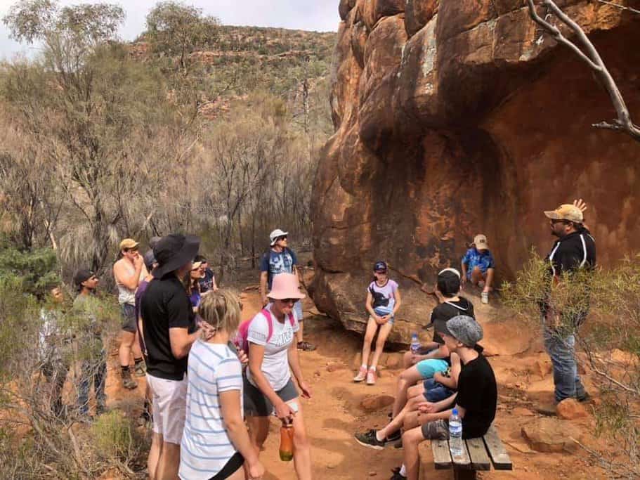 A group of people, including children and adults, gather at a significant cultural site in the Flinders Ranges, listening to a guide explain the ancient rock art and heritage. This image highlights the immersive experience of the Flinders Ranges Tour, showcasing the educational and cultural aspects of the journey.