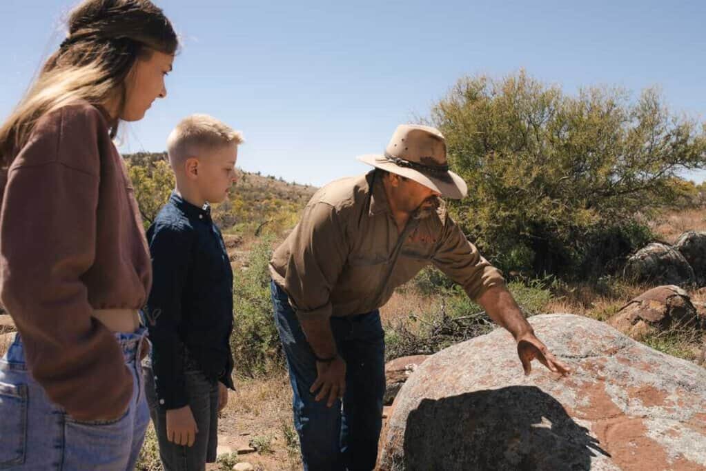 A guide points out ancient markings on a rock to two children during a Flinders Ranges Tour, offering an educational experience about the Adnyamathanha culture and their historical sites. This image illustrates the interactive and informative nature of the tour, highlighting its appeal to families.
