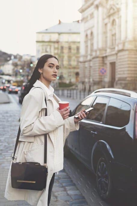 Woman in a stylish coat waiting on a busy street, checking her smartphone possibly for an Uber ride in Melbourne.