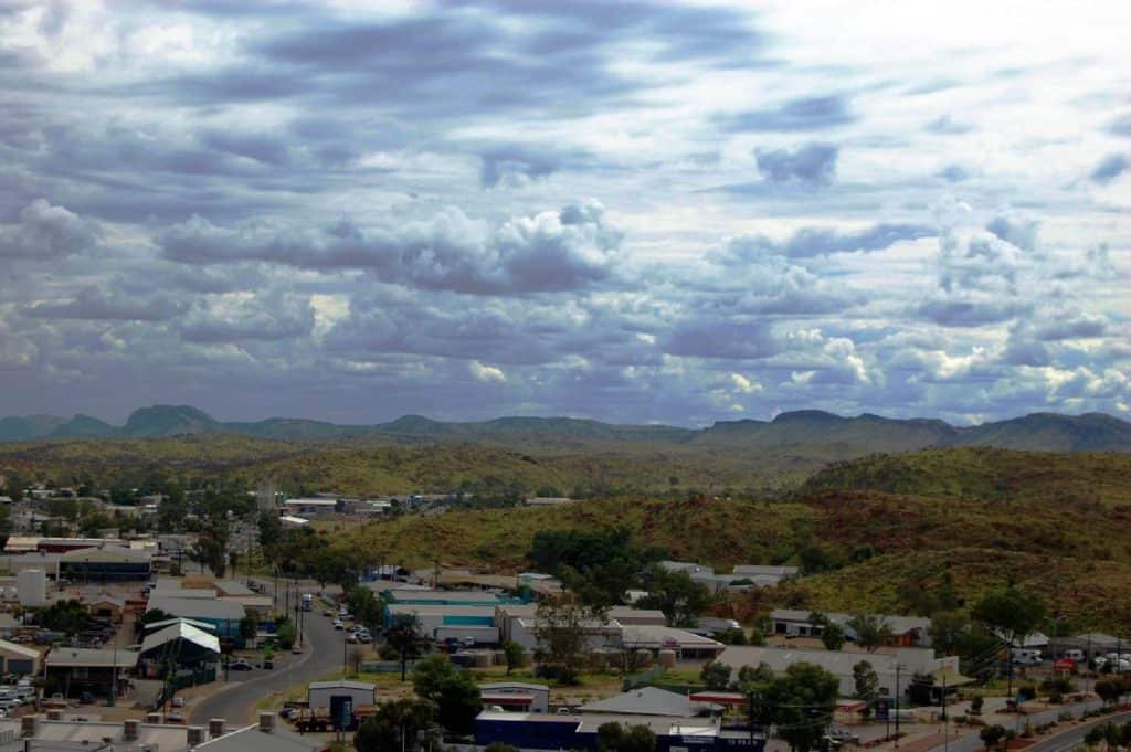 A panoramic view of Alice Springs, showing a mix of buildings, roads, and surrounding hills under a cloudy sky. The town’s remote location and small-community feel are key factors when weighing the advantages and disadvantages of living in Alice Springs.