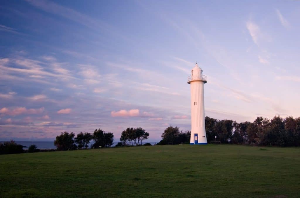 Yamba Lighthouse standing tall on a grassy headland under a pastel sunset sky, a peaceful lookout along the scenic road trip between Sydney and Byron Bay.