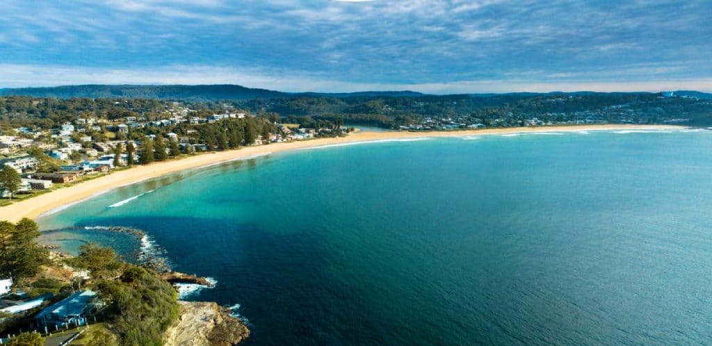 Aerial view of Avoca Beach curving along the Central Coast, framed by calm turquoise waters and bushland—an ideal first stop on a road trip between Sydney and Byron Bay.