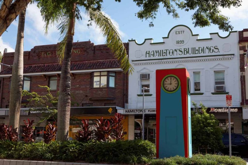A colourful clock sculpture in Mackay's city centre with historic buildings and palm trees in the background, representing the lifestyle and amenities discussed in the post about the pros and cons of living in Mackay.