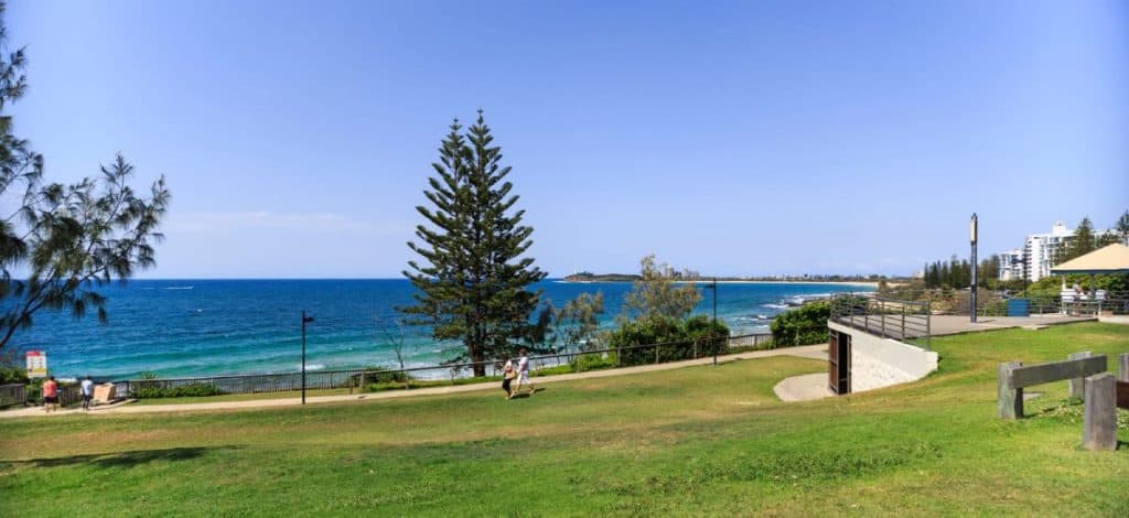 People walking along a coastal path overlooking the ocean at Mooloolaba on a sunny day, with grassy parklands, Norfolk pine trees, and apartment buildings in view – a typical outdoor lifestyle scene that highlights the pros and cons of living in Sunshine Coast, especially the appeal of nature and beachside living.