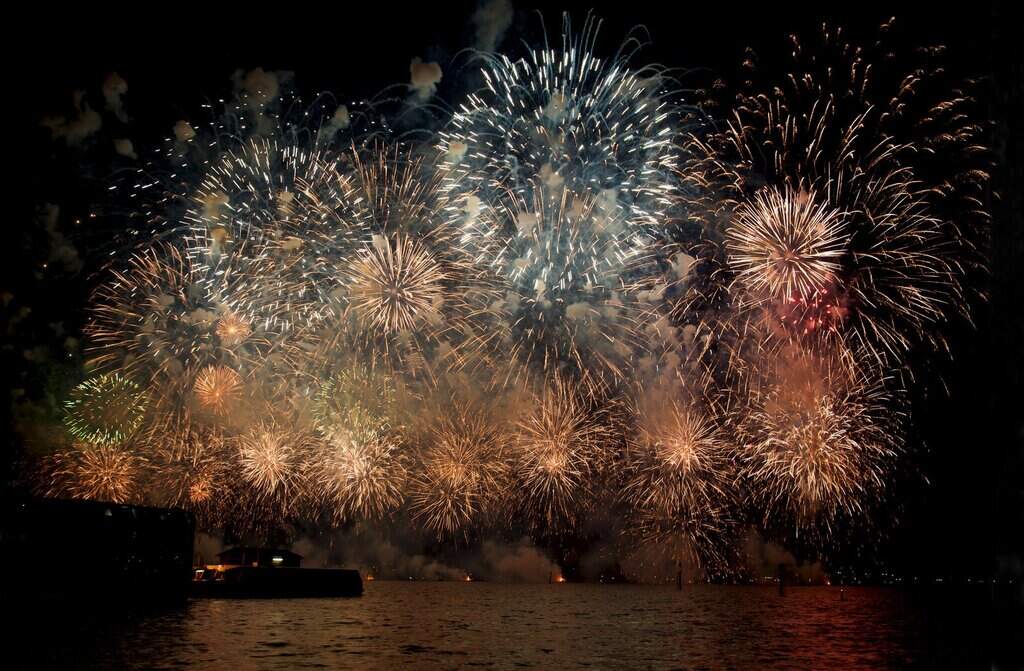 Fireworks explode in a dazzling array of gold, white, and subtle greens over the Swan River during Australia Day in Perth. Silhouetted boats and structures line the water as the night sky fills with smoke and sparkling light.