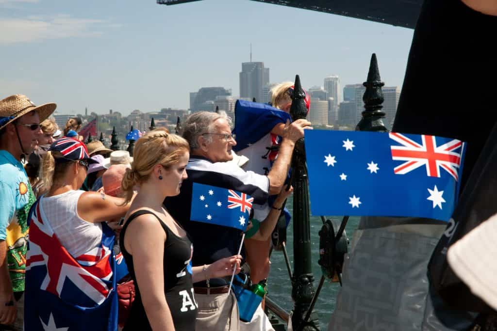 Crowd of people gathered along the waterfront under the Sydney Harbour Bridge, waving Australian flags and dressed in festive attire. The image captures a lively celebration of Australia Day in Sydney, with city buildings visible in the background and a sense of national pride throughout.