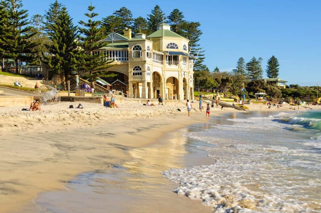 People relax and stroll along the sandy shore of Cottesloe Beach under clear blue skies on Perth's Australia Day. The iconic art deco-style Indiana Tea House overlooks gentle waves and sunlit sand, framed by tall pine trees and coastal greenery.