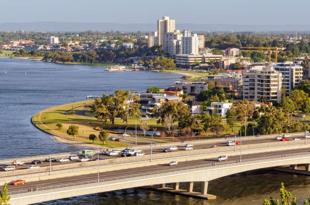 A view of the Perth cityscape along the Swan River shows cars crossing a busy bridge while the riverside park curves around the water. High-rise buildings and leafy neighborhoods reflect the relaxed yet vibrant atmosphere typical of Australia Day celebrations in Western Australia.