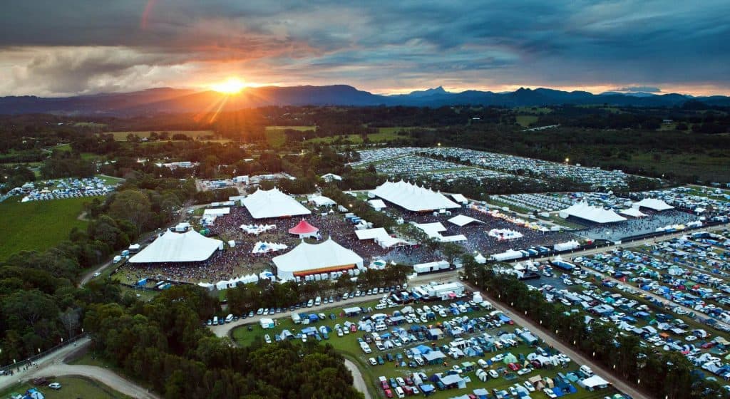 Aerial view of a large outdoor music festival at sunset with multiple white stage tents, packed crowds, and rows of camping tents and parked cars, showing the scale and atmosphere of Bluesfest, one of the best music festivals in Australia