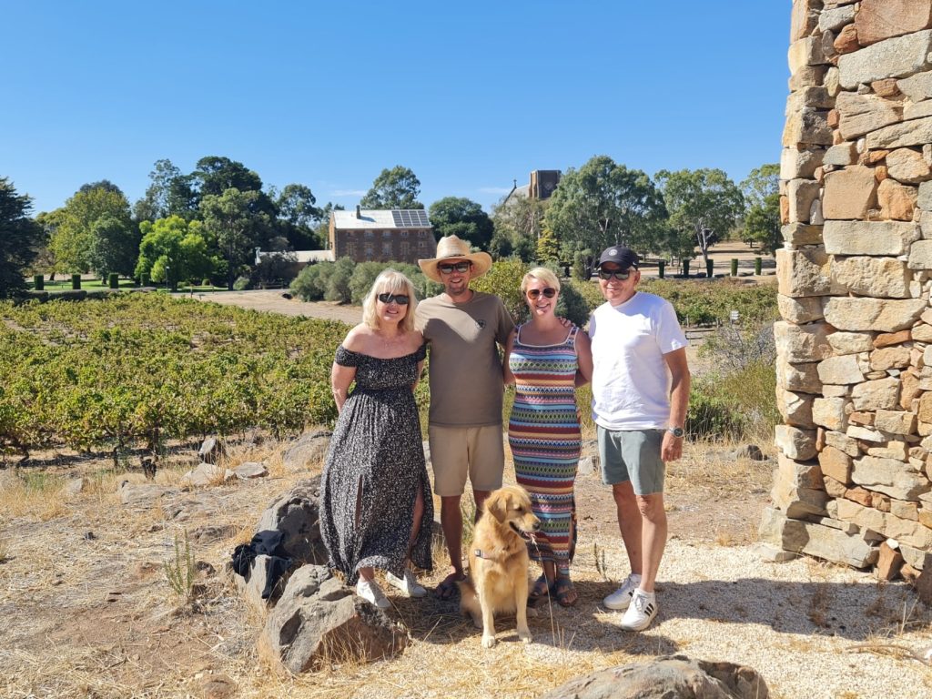 Group of friends with their dog standing in front of historic buildings and vineyards in Clare Valley, highlighting the scenic stops and savings available with a clare valley tour discount code.