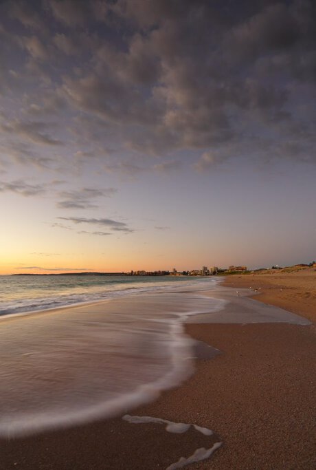 Golden sunset over a wide sandy beach with gentle waves washing onto the shore and a distant coastal skyline, showcasing one of Australia’s Best Beaches and the relaxed seaside beauty featured in the blog post.