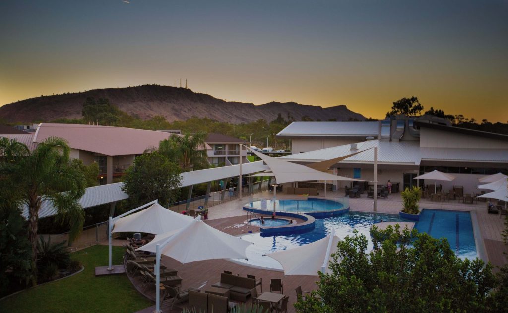 Resort-style hotel in Alice Springs with a large swimming pool, shaded sun loungers, and views of the MacDonnell Ranges at sunset, highlighting a top-rated resort option for travellers deciding where to stay in Alice Springs for a relaxing outback getaway.