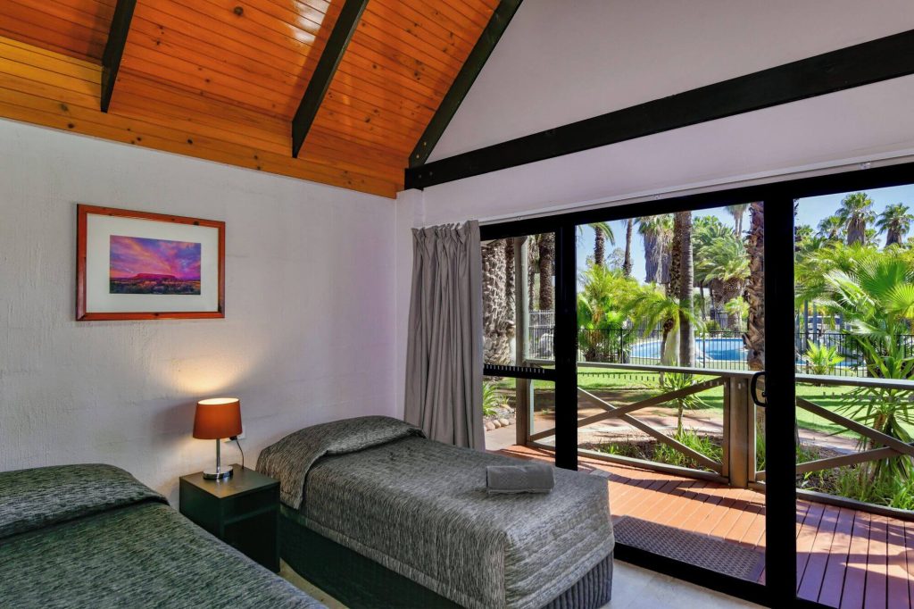 Villa-style accommodation in Alice Springs with twin beds, timber ceiling, and sliding doors opening to lush gardens and a swimming pool, perfect for travellers researching family-friendly accommodation in Alice Springs Australia.