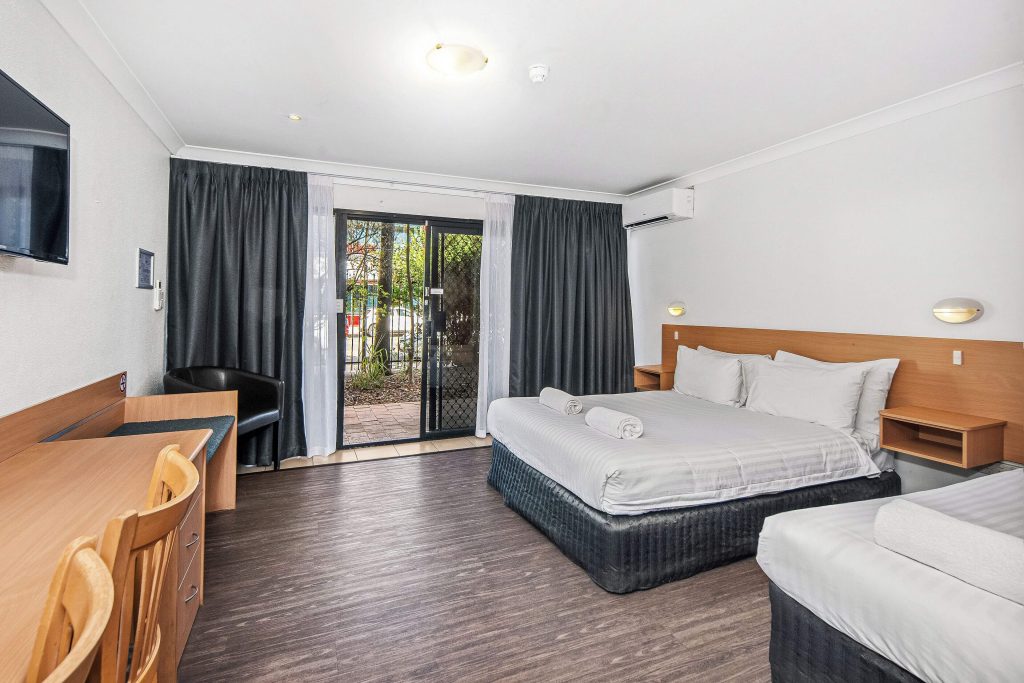 Spacious budget hotel room in Alice Springs with a queen and single bed, work desk, TV, and sliding door to a private courtyard, a practical choice for travellers comparing cheap accommodation in Alice Springs NT.