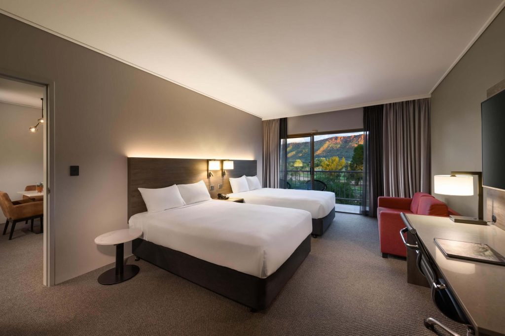 Spacious hotel room in Alice Springs with two double beds, desk, red lounge chair, and balcony overlooking the MacDonnell Ranges, a great option for families comparing hotels in Alice Springs NT and deciding where to stay in Alice Springs.