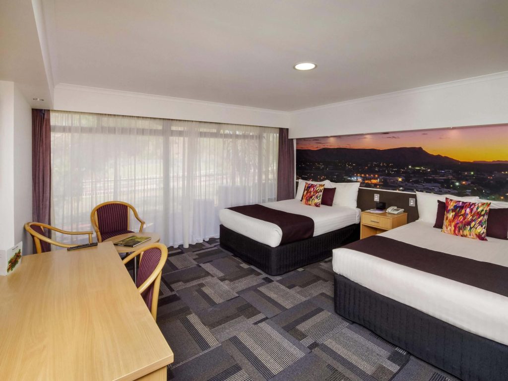 Spacious twin hotel room in Alice Springs with two double beds, desk, seating area, and large window with sunset views over the MacDonnell Ranges, perfect for families searching for the best accommodation in Alice Springs NT.