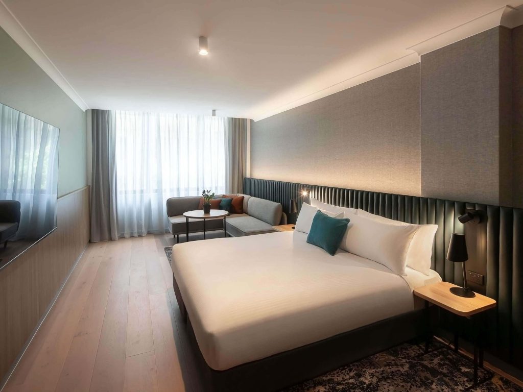 A stylish king room at Novotel Sydney City Centre, a top mid-range hotel in the Sydney CBD, featuring a plush king bed, teal velvet headboard, timber floors, and a soft lounge area.