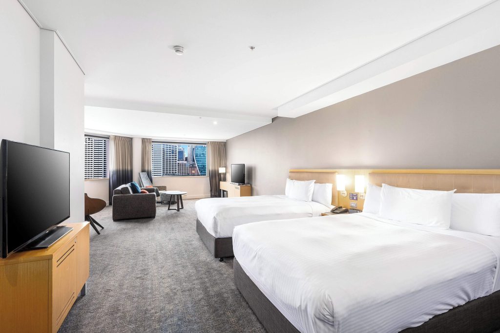 A spacious twin room at Novotel Sydney on Darling Harbour, one of the best family-friendly hotels in Sydney, featuring two queen beds, city skyline views, and a comfortable lounge area.