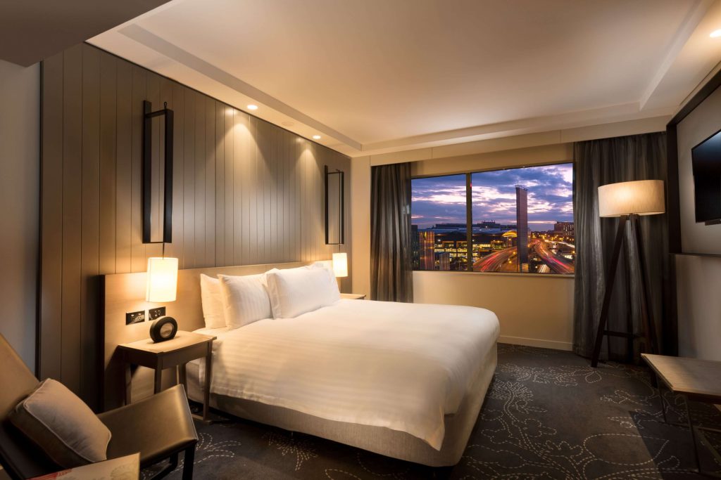 Modern hotel room with large bed and window overlooking Darling Harbour and Sydney city lights at sunset, showcasing comfortable Darling Harbour hotels in one of the best areas to stay in Sydney for tourists