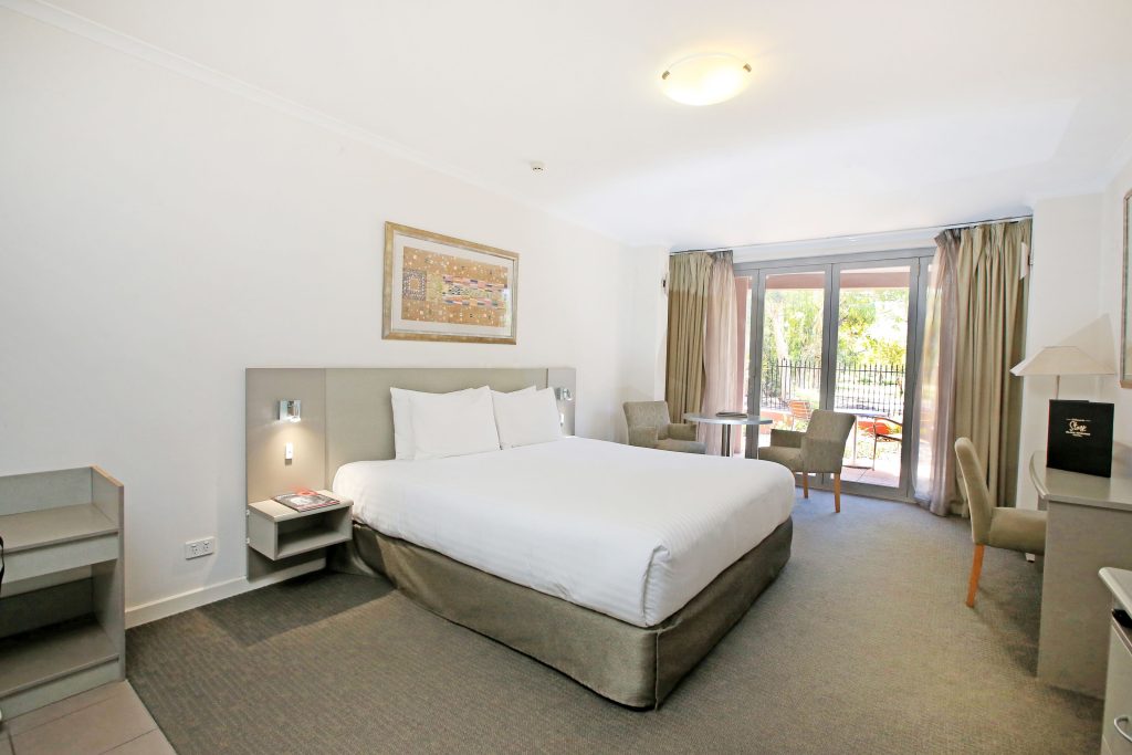 Modern hotel room in Alice Springs with a king bed, bedside tables, desk, armchairs, and balcony doors overlooking greenery, showcasing a comfortable option for travellers researching Where to Stay in Alice Springs.