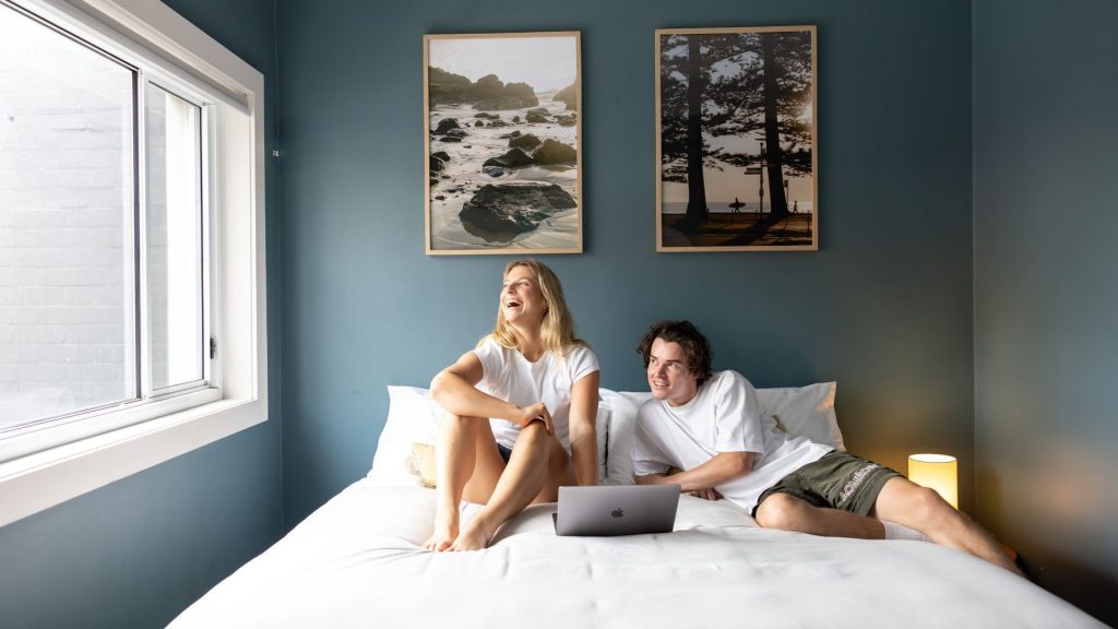 Young travellers relaxing on a bed with laptop at Stoke Beach House in Manly, highlighting budget-friendly Manly Sydney accommodation in one of the best suburbs to stay in Sydney near the beach
