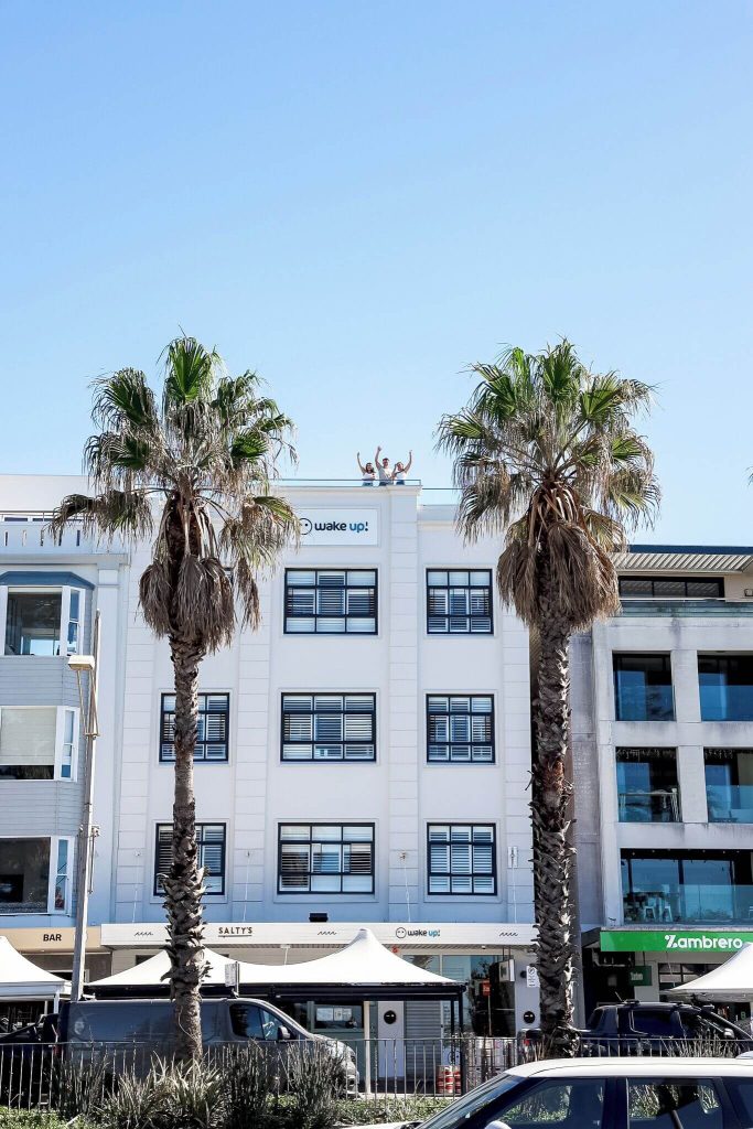 Wake Up! hostel building framed by palm trees along Bondi Beach, showcasing popular Bondi Beach accommodation and a great option for travellers choosing the best suburbs to stay in Sydney near the coast