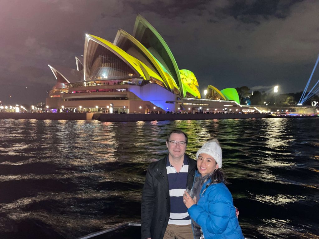 Couple enjoying harbour views with Sydney Opera House lit in vibrant colours at night, capturing the iconic light projections and atmosphere of Vivid Sydney in 2023.