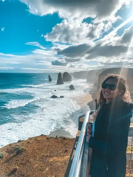 Traveler wearing sunglasses and a dark coat stands at a lookout along the Great Ocean Road, overlooking the Twelve Apostles sea stacks and rugged coastline. Sunlight breaks through dramatic clouds above turquoise waves and sandy beaches, highlighting one of the most iconic stops featured in a Great Ocean Road ultimate guide.