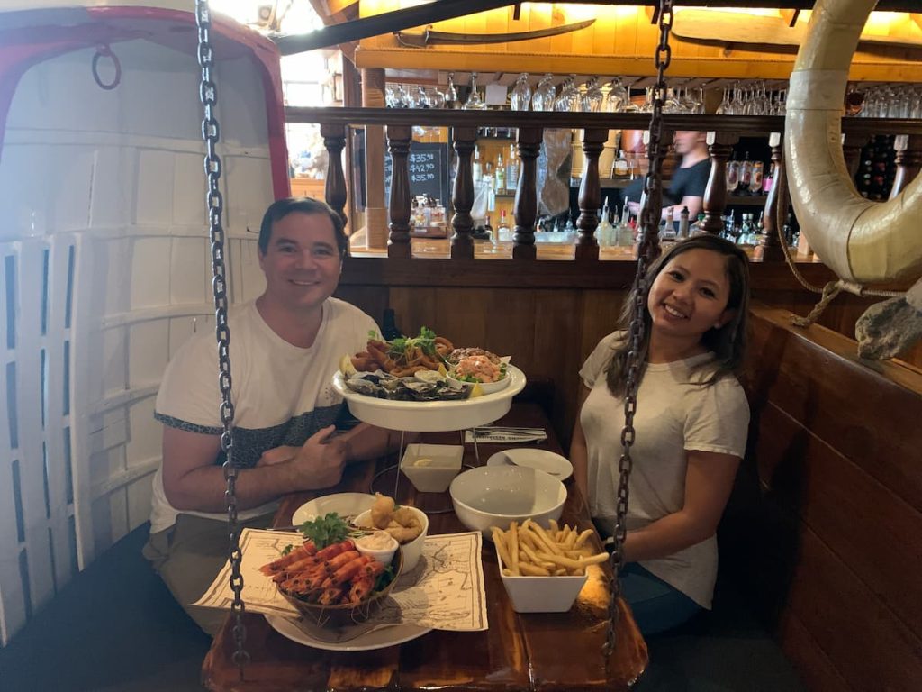 Two diners seated inside Drunken Admiral in Hobart with a large seafood platter, prawns, oysters, fries, and other seafood dishes served on a rustic wooden table surrounded by nautical decor. A popular dining stop on a Tasmania itinerary, this waterfront restaurant is known for its maritime atmosphere and fresh Tasmanian seafood.