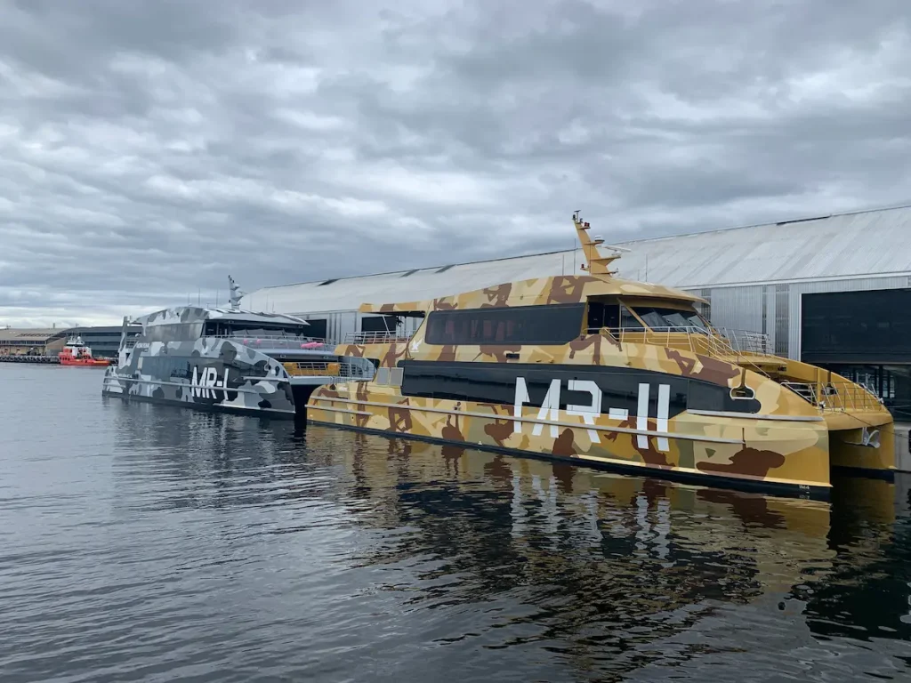 Camouflage patterned MONA ferry vessels docked at Hobart's waterfront beneath an overcast sky, with their reflections visible on the calm harbor water. A memorable part of a 9 day Tasmania itinerary, the ferry ride to MONA offers a unique journey along the Derwent River before arriving at one of Tasmania's most famous cultural attractions.