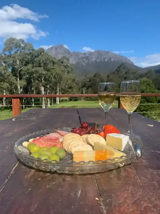 Cheese board with crackers, olives, cured meats, fruit, and sparkling wine on an outdoor table overlooking mountain views near Sheffield, Tasmania. A memorable stay on a Tasmania road trip itinerary, this countryside accommodation combines local produce, peaceful surroundings, and stunning views of the Cradle Mountain region.