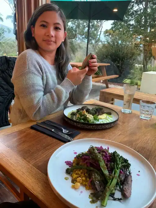 Person seated at a table enjoying a meal at On the Bay Restaurant in Coles Bay, with plates of locally inspired dishes and views of native bushland through large windows. A delicious stop on a Tasmania food and travel itinerary, this waterfront dining experience offers a chance to enjoy fresh local produce near Freycinet National Park.