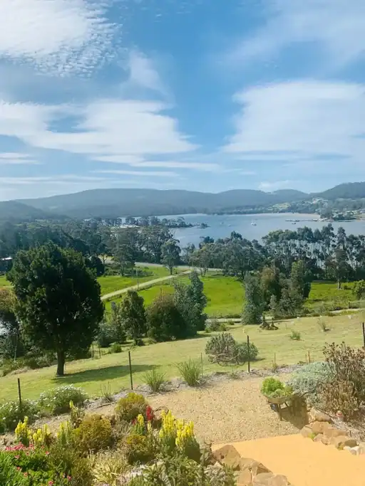 View from the balcony at Storm Bay Bed and Breakfast in Port Arthur overlooking rolling green countryside, gardens, and a sheltered bay surrounded by forested hills beneath a bright blue sky. A relaxing accommodation stop on a Tasmania itinerary, this peaceful setting offers scenic water views and easy access to the natural attractions of the Tasman Peninsula.