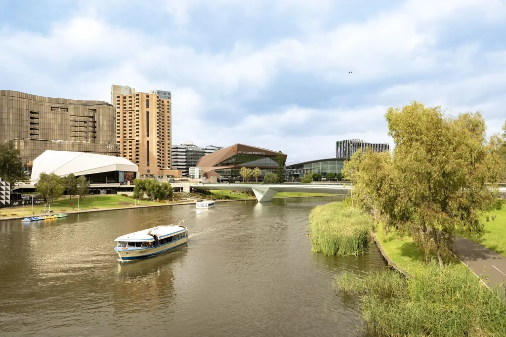 Boats cruise along the River Torrens with the Adelaide skyline and Convention Centre in the background. A riverside walk is one of the top Adelaide attractions in the city center.