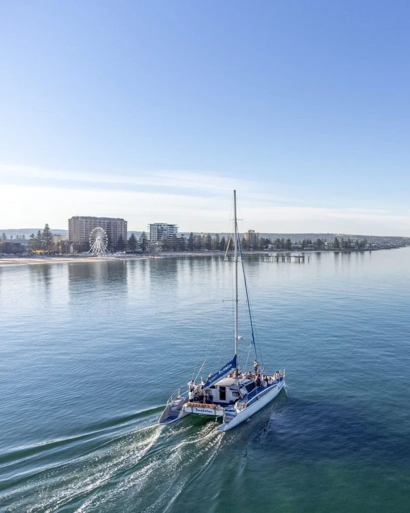A catamaran cruises across the calm waters off Glenelg with the Adelaide coastline in the background. A sunset cruise is one of the best ways to experience Adelaide travel from the water.