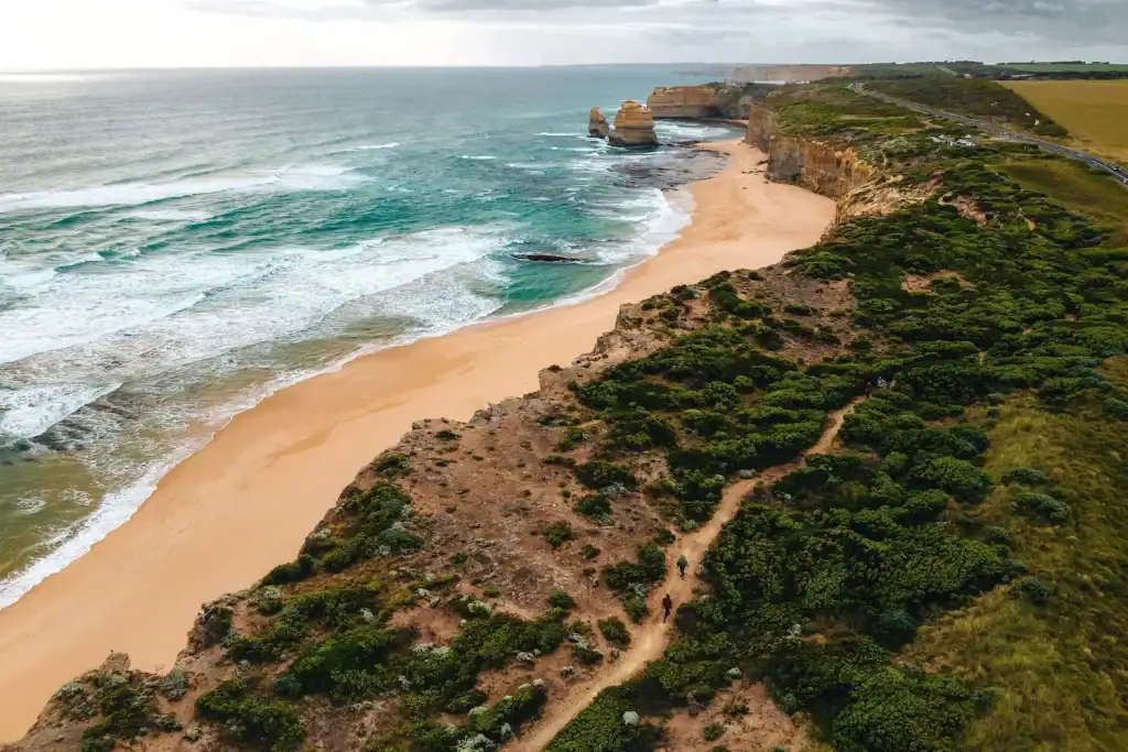 Aerial view of hikers following the Great Ocean Walk above golden beaches and rugged limestone cliffs with the Twelve Apostles in the distance. This spectacular stretch of Victoria's coastline is one of the highlights of an Australia hiking guide.
