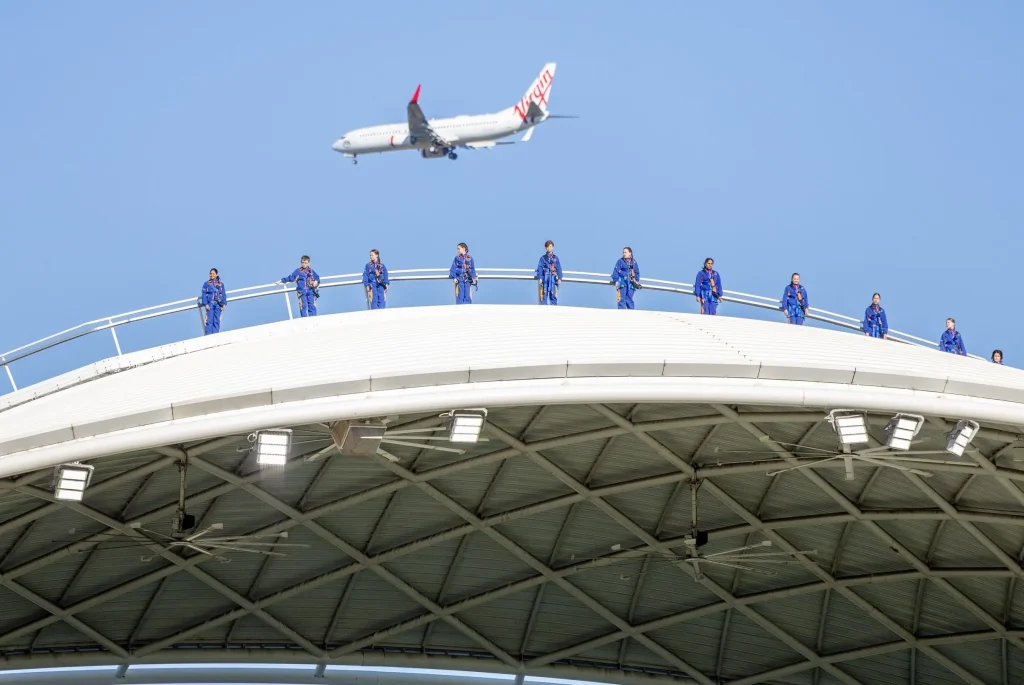 Visitors wearing blue jumpsuits walk across the curved roof of Adelaide Oval while a Virgin Australia plane flies overhead. RoofClimb is one of the most unique Adelaide attractions for adventurous visitors.
