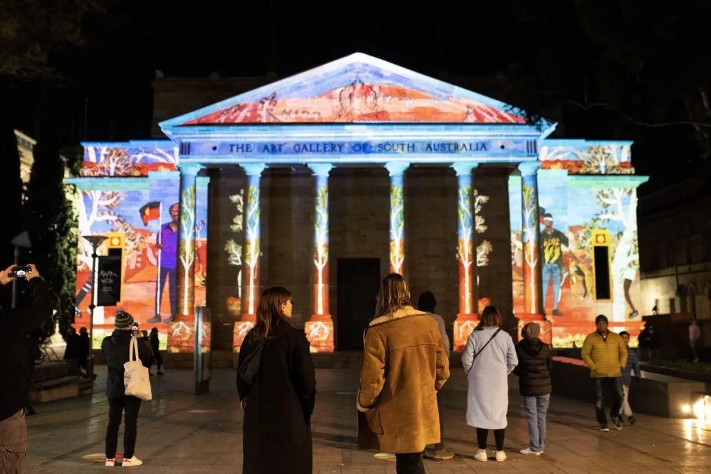 Visitors watch colorful light projections illuminate the Art Gallery of South Australia during a nighttime festival. This seasonal display is one of the best things to do in Adelaide after dark.
