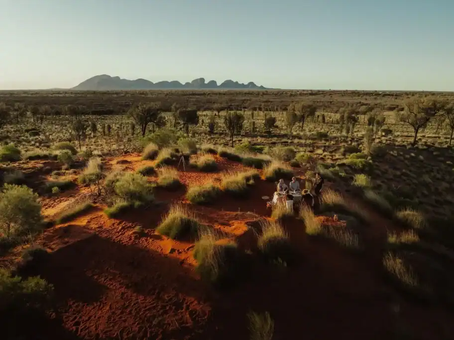 Aerial view of visitors dining in the Australian outback surrounded by red sand, native grasses, and Kata Tjuta on the horizon. The remote desert landscape highlights the dramatic scenery found along Great Walks Australia.