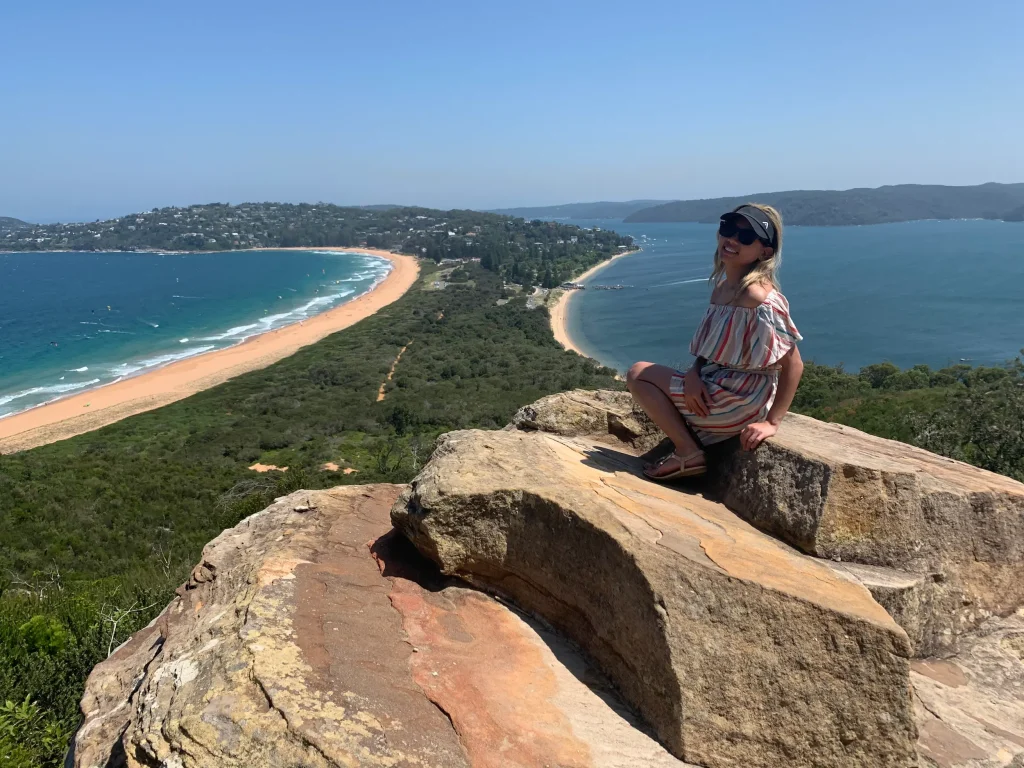A traveler sits on a sandstone rock near Barrenjoey Lighthouse, overlooking the golden sands of Palm Beach and the calm waters of Pittwater on a clear, sunny day. Visiting Barrenjoey Head is one of the best things to do in Sydney, with panoramic coastal views and one of the city's most iconic lighthouse walks.