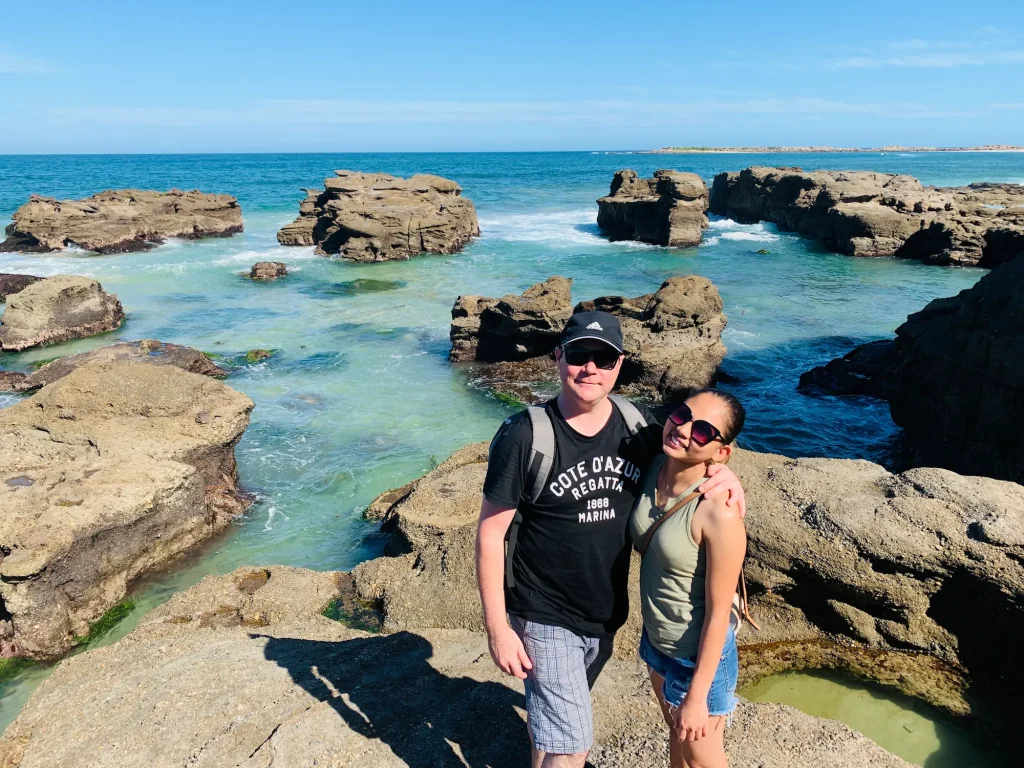 A couple stands on the rocky shoreline at Caves Beach, surrounded by crystal-clear turquoise water, rugged sandstone formations, and the open ocean under a bright blue sky. Visiting Caves Beach is one of the most memorable coastal day trips from Sydney, known for its sea caves, rock pools, and stunning shoreline along the Lake Macquarie coast.