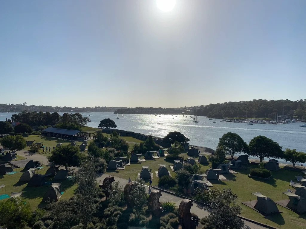 An elevated view of Cockatoo Island shows rows of camping tents overlooking Sydney Harbour, where sailboats and yachts drift across the sparkling water under a clear blue sky. Camping on Cockatoo Island is one of the most unique things to do in Sydney, combining waterfront views with the island's rich maritime and convict history.