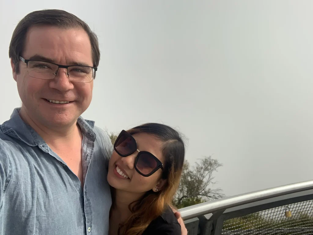 A couple smiles for a selfie at Echo Point Lookout in the Blue Mountains, standing beside the viewing platform railing with misty skies and treetops in the background. Visiting Echo Point is one of the best places to visit outside Sydney, offering breathtaking escarpment views and easy access to the region's iconic walking trails.