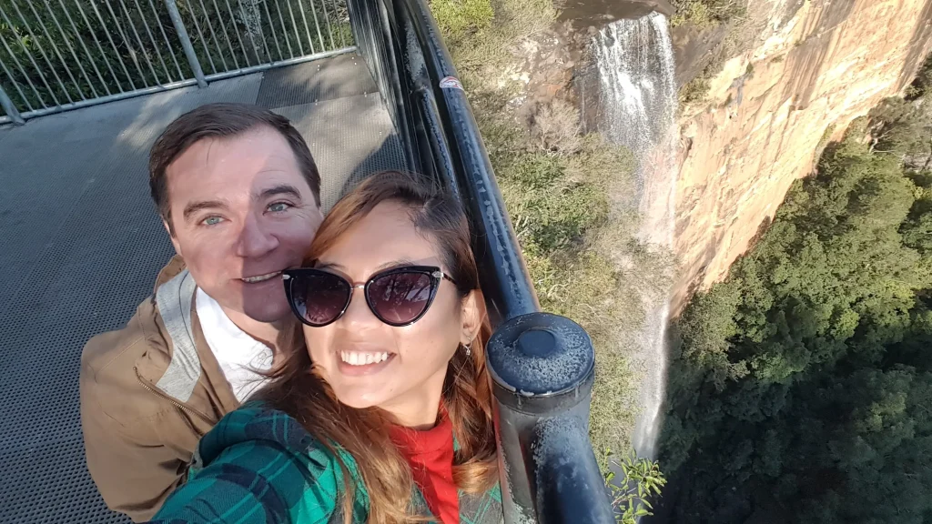 A couple takes a selfie from the Fitzroy Falls lookout platform with the towering waterfall cascading over sandstone cliffs into the lush valley below. Visiting Fitzroy Falls is one of the best day trips from Sydney, offering spectacular waterfall views and scenic walking trails in the Southern Highlands.