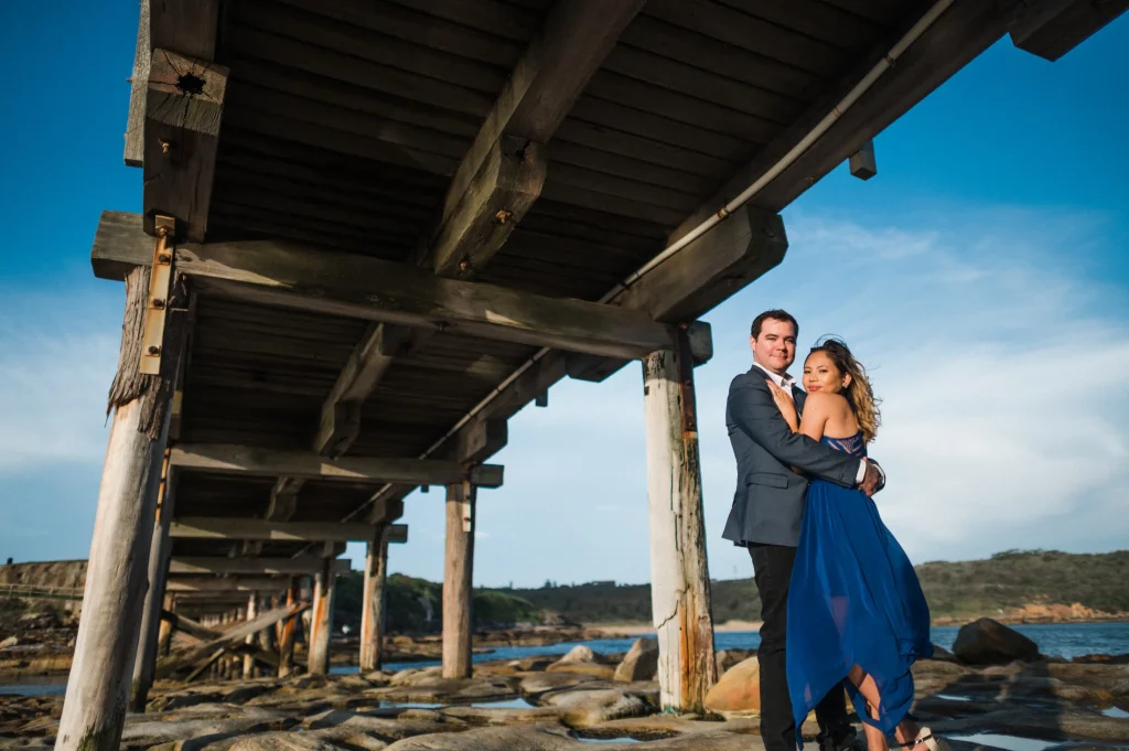 A couple embraces beneath the historic wooden jetty at La Perouse, standing on a rocky shoreline with calm coastal water and a clear blue sky in the background. This picturesque waterfront spot is one of the most romantic places to visit in Sydney, offering beautiful coastal scenery and a peaceful escape by the harbour.